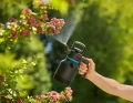 Opryskiwacz ciśnieniowy 1,25 l Gardena z czarną dyszą. Uchwyt z pomarańczowym przyciskiem. Trzymany pionowo na tle drzew, używany w ogrodzie.