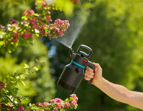 Opryskiwacz ciśnieniowy 1,25 l Gardena z czarną dyszą. Uchwyt z pomarańczowym przyciskiem. Trzymany pionowo na tle drzew, używany w ogrodzie.