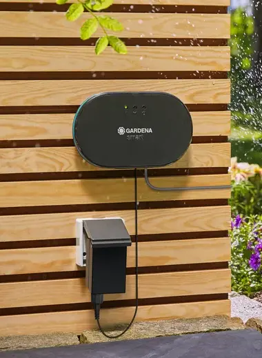 Smart sterownik nawadniania Gardena, w kolorze czarnym, widok zamontowanego na płycie sterownika wraz z ładowarką i przewodami.