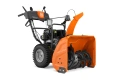 Odśnieżarka ST269 Husqvarna w kolorze pomarańczowym, frontalnie zmontowana, z metalową, szeroką turbiną do odśnieżania. Widoczne gumowe koła oraz ergonomiczne uchwyty. Silnik spalinowy, z czytelnym logo Husqvarna na uchwycie.