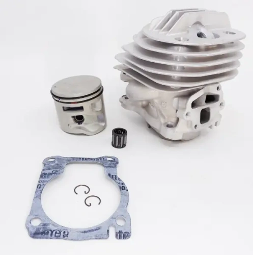 Cylinder kompletny pilarki Husqvarna 564 XP w kolorze szarym z charakterystycznym żeberkowaniem na obudowie. Widoczny metalowy tłok w srebrnym kolorze obok cylindra, wraz z pierścieniem oraz elementem montażowym. Produkt leży na jasnym tle.