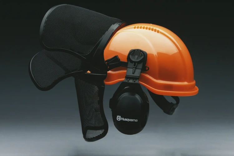 Kask leśny Functional Husqvarna Orange, ujęcie boczne, siatkowa osłona uniesiona do góry, widoczny napis Husqvarna. Tło jednolite.