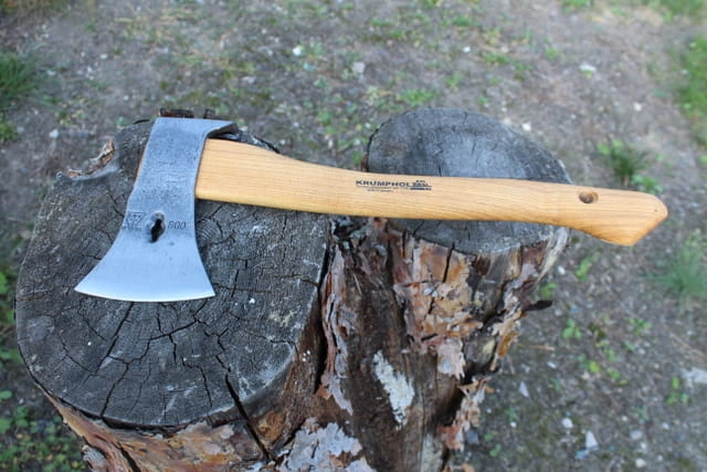 Siekiera ciesielska Krumpholz ważąca 0.8 kg z drewnianym trzonkiem. Ujęta z boku na pniu drzewa, z wyraźnym logo. Ostra, metalowa głowica o prostym kształcie, szlifowana. Naturalne tło, z widocznymi detalami słojów drewna.