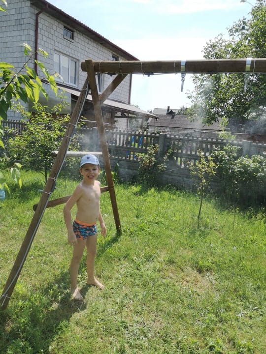 Kurtyna wodna Gardena w ogrodzie, zamontowana w górnej części huśtawki. Dziecko w kąpielówkach stoi na trawie, ciesząc się mgiełką z wodą. Jasne niebo w tle, drzewa i ogrodowe płoty tworzą typowy letni krajobraz.