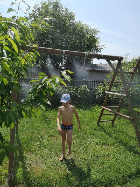 Kurtyna wodna Gardena zamontowana nad huśtawką w ogrodzie, w otoczeniu zieleni. Dziecko w kąpielówkach i czapce stoi w strefie rozpylania wody, na tle drzewa i drewnianego ogrodzenia, w świetle słonecznym.