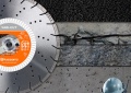 Tarcza diamentowa Husqvarna Vari-Cut o średnicy 400 mm i otworze 25,4 mm, z wyraźnym logo na pomarańczowym tle. Ujęcie frontalne, tarcza leży na podłożu, w tle różne materiały budowlane, podkreślające zastosowanie narzędzia w cięciu.