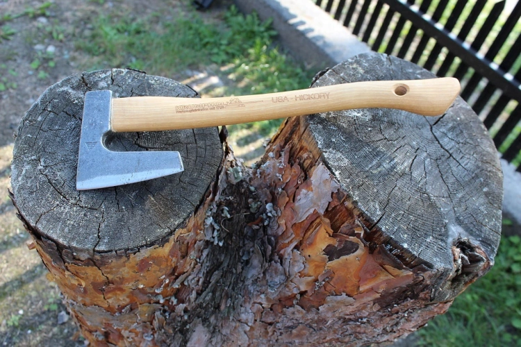 Toporek myśliwski Krumpholz z drewnianą rączką w kolorze jasnego hikorowego drewna, metalową głowicą w kształcie litery L. Umieszczony na ciemnym, gładkim kłodzie. Widoczne logo Krumpholz i USA-Hickory. Ujęcie w zbliżeniu.